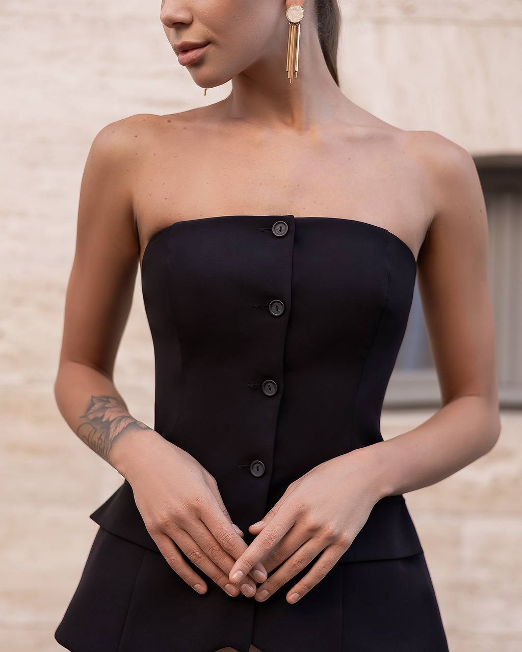 A woman wearing a black strapless corset-style dress with a front button placket and peplum hem, shown from the shoulders to the hips against a light beige stone wall.