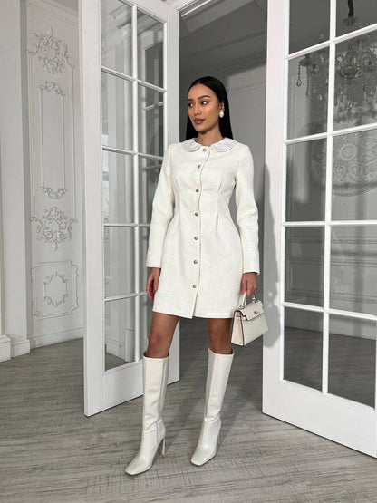 Woman in a white coat and knee-high boots standing in a room with large windows.
