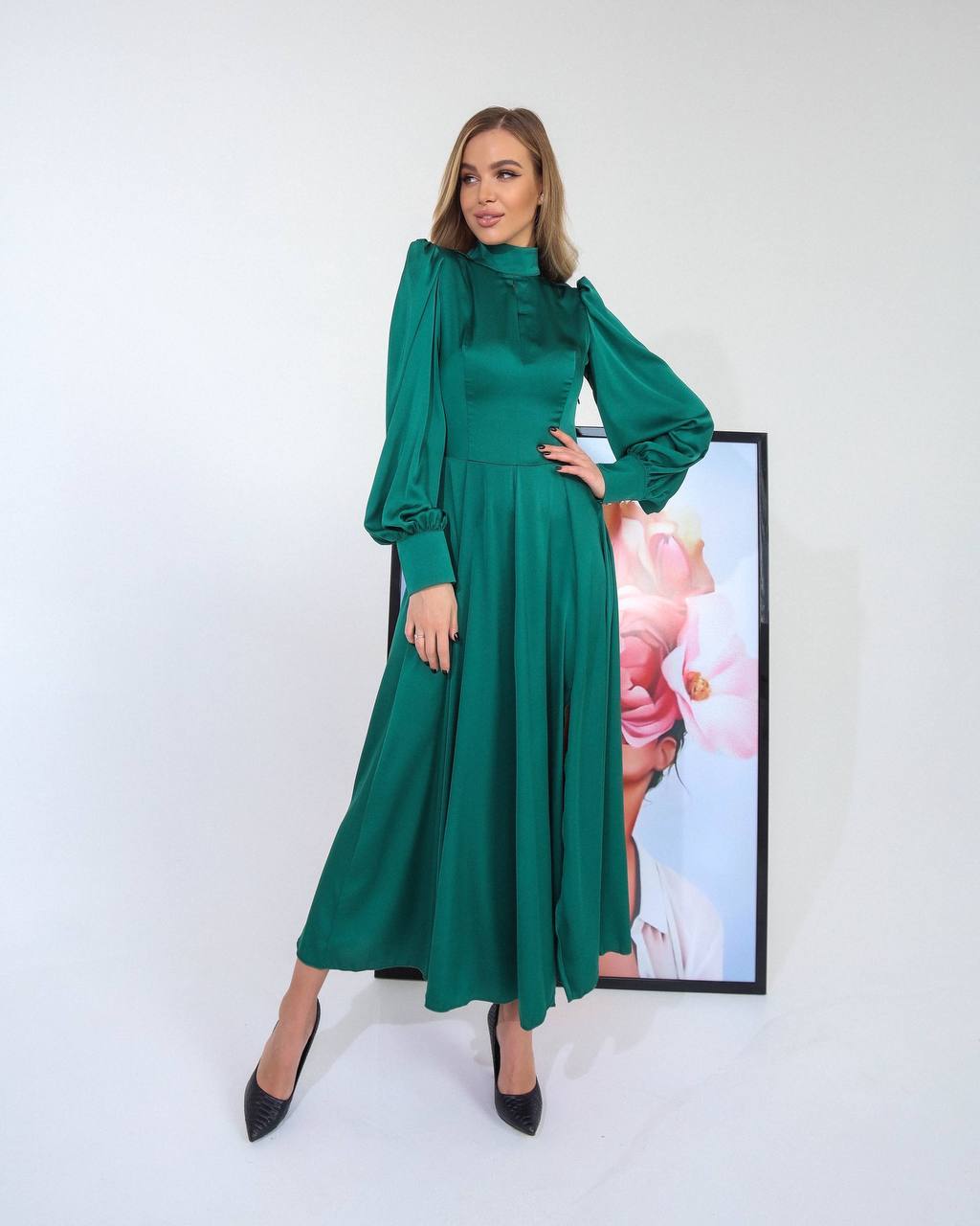 Woman wearing a green dress standing against a white background with a framed picture of flowers in the background.
