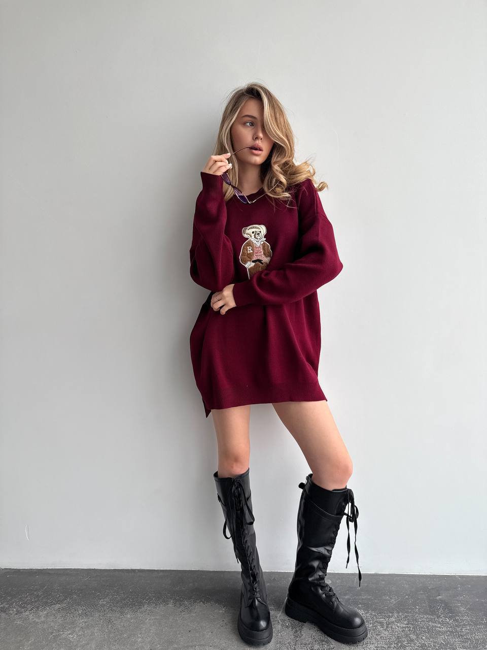 Woman wearing a burgundy sweater dress with black knee-high boots against a plain background