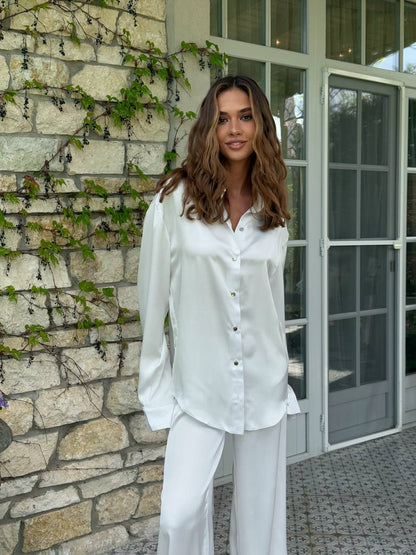 Woman in a white outfit standing in front of a stone wall with greenery.