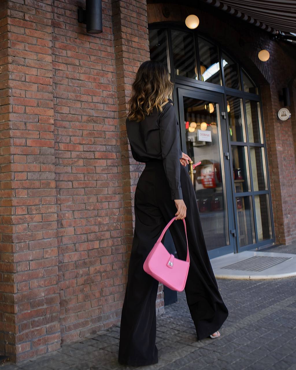 Woman in black outfit with pink handbag walking past a brick building.
