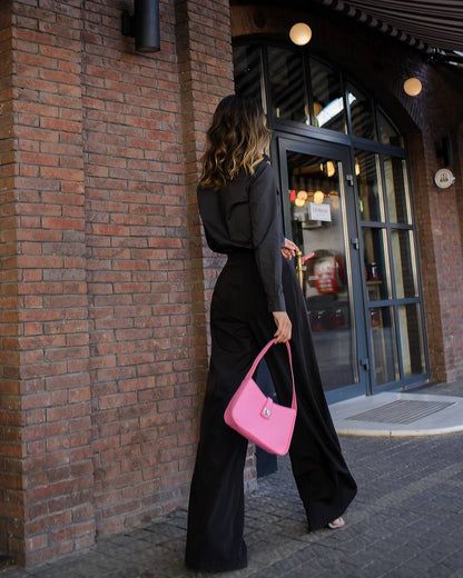 Woman in black outfit with pink handbag walking past a brick building.