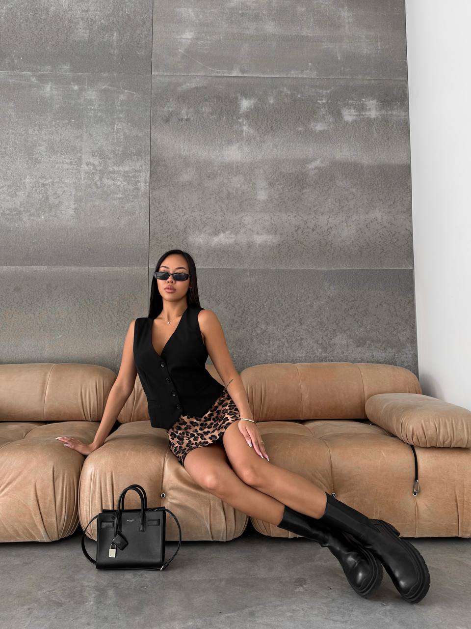 Woman sitting on a beige sofa with a black handbag in front of a gray wall.