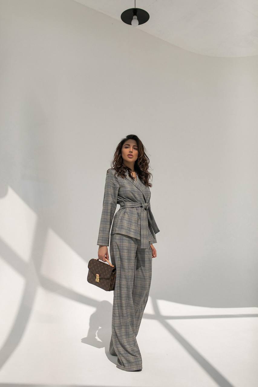Woman in a gray checkered suit standing against a white wall with a minimalistic background.