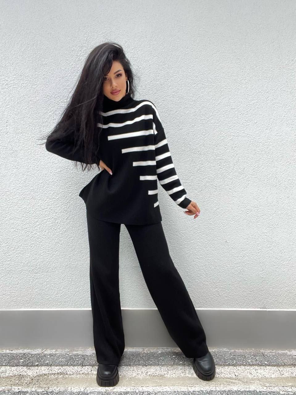 Person wearing a black and white striped sweater and black pants against a light gray wall.