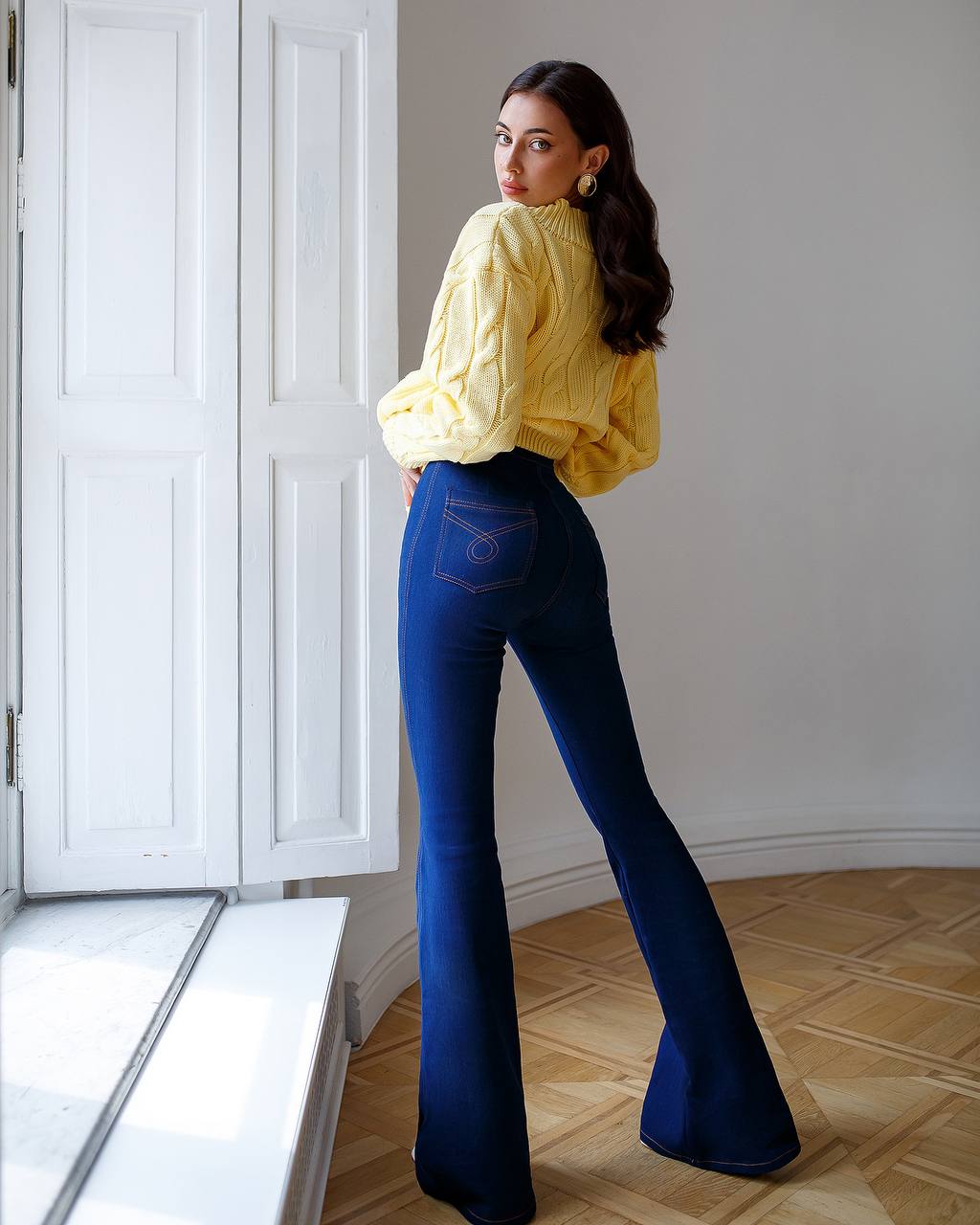 Woman wearing a yellow sweater and blue flared jeans standing in a room with white walls and wooden floor.