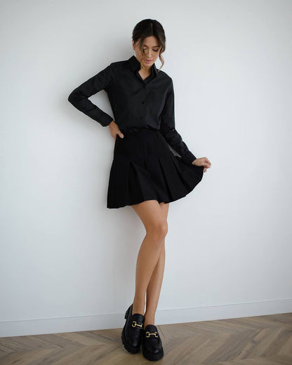 Woman wearing a black shirt and skirt with black shoes against a white wall.