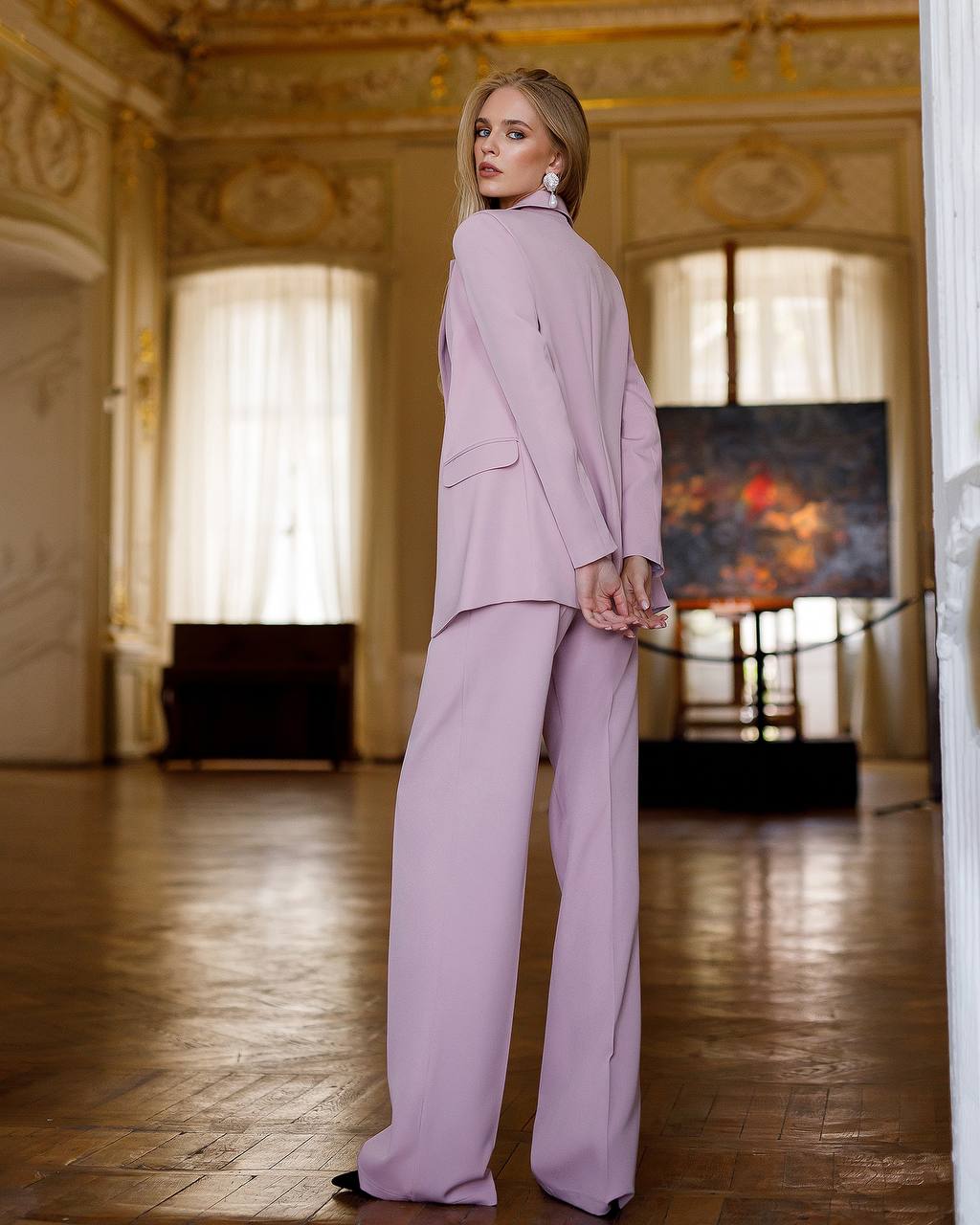 Woman in a lavender suit standing in an elegant room with wooden floor and decorative walls.