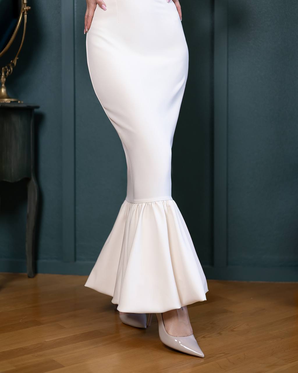 White mermaid-style dress with a ruffled bottom on a wooden floor against a dark blue wall.