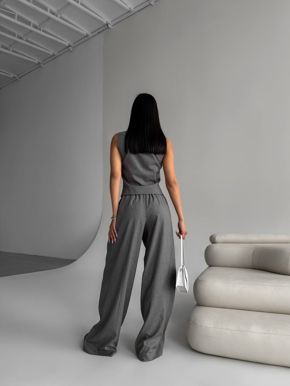 Woman in a gray jumpsuit standing in a minimalistic room with a modern sofa.