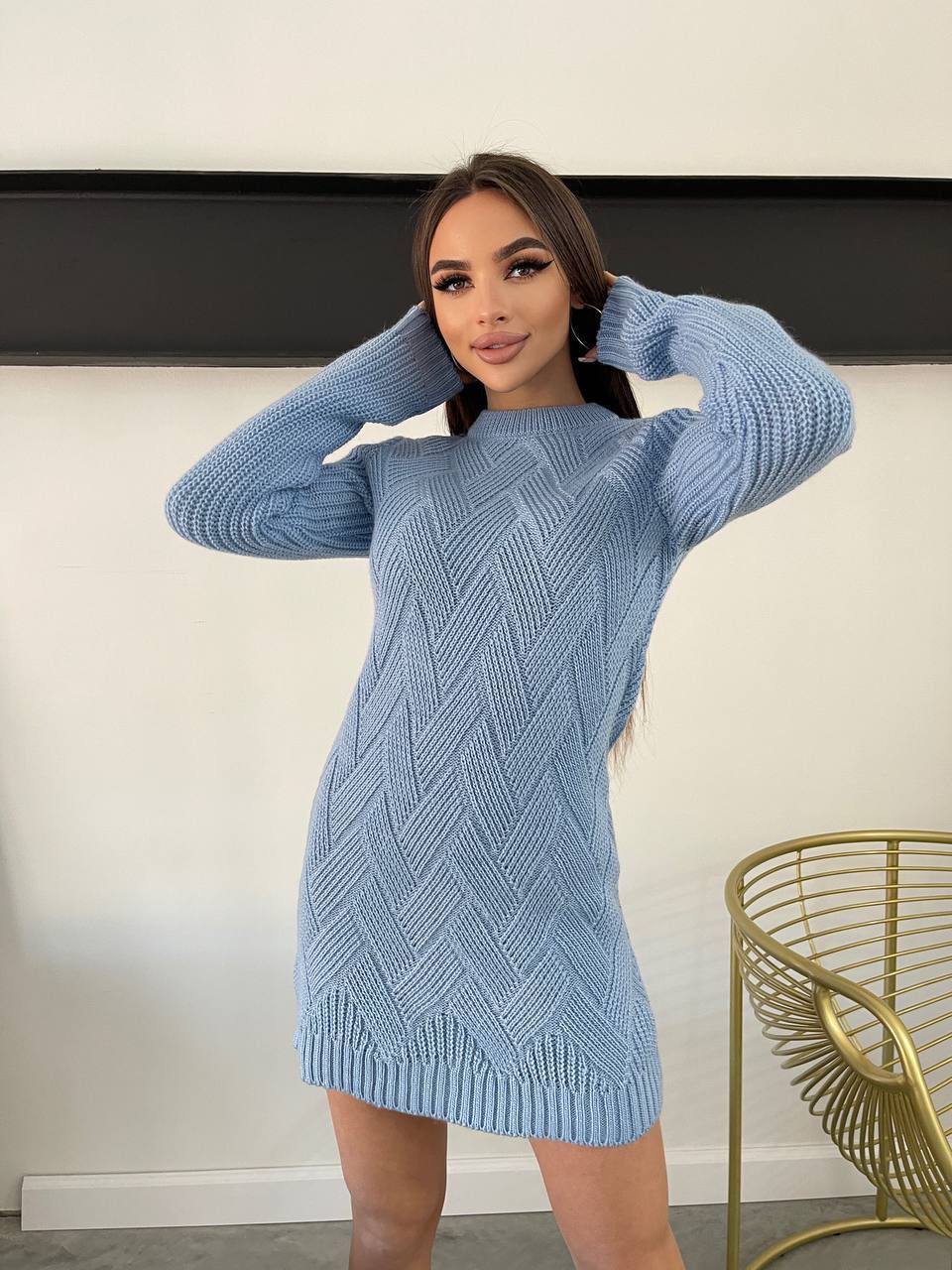 Woman wearing a blue knitted dress with a white wall and gold chair in the background