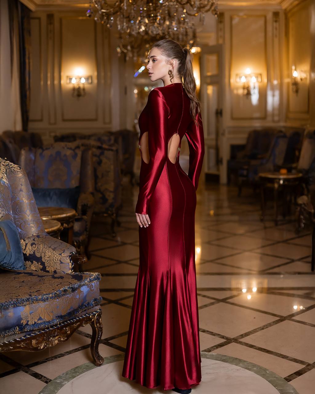 Woman in a red evening gown standing in an elegant room with chandeliers and furniture.