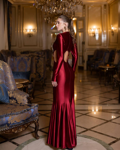 Woman in a red evening gown standing in an elegant room with chandeliers and furniture.