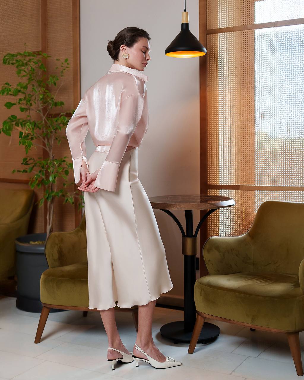 Woman in a white outfit standing in a modern interior setting with green chairs and a lamp.