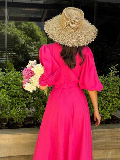 Woman in a bright pink dress and straw hat holding flowers, standing outdoors.
