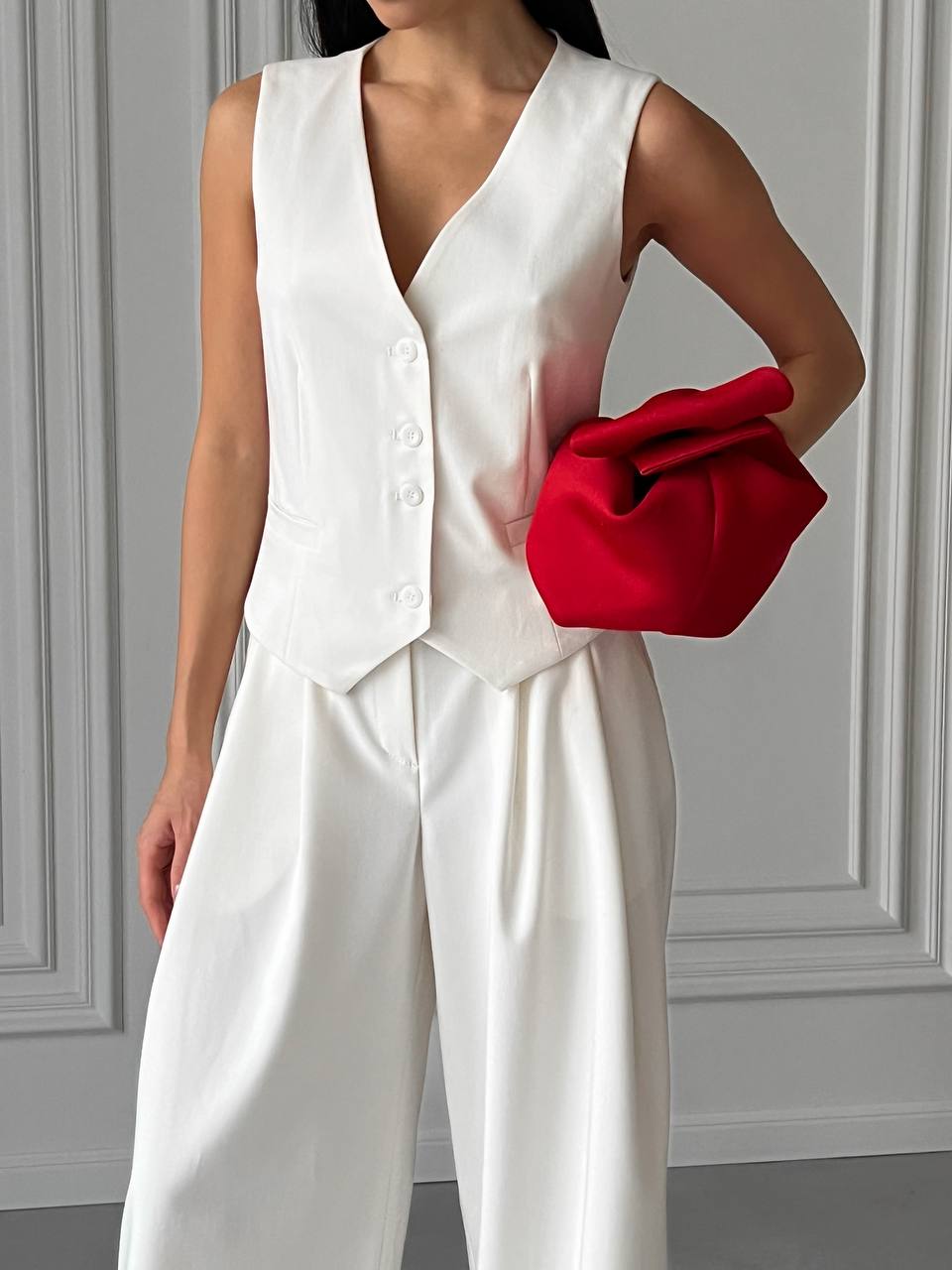 Person wearing a white outfit holding a red clutch against a gray wall.