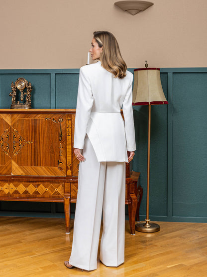 A woman stands with her back to the camera wearing a white tailored suit with a structured jacket and wide-leg trousers in an indoor room with wood floors, teal wainscoting, a wooden cabinet, and a floor lamp.