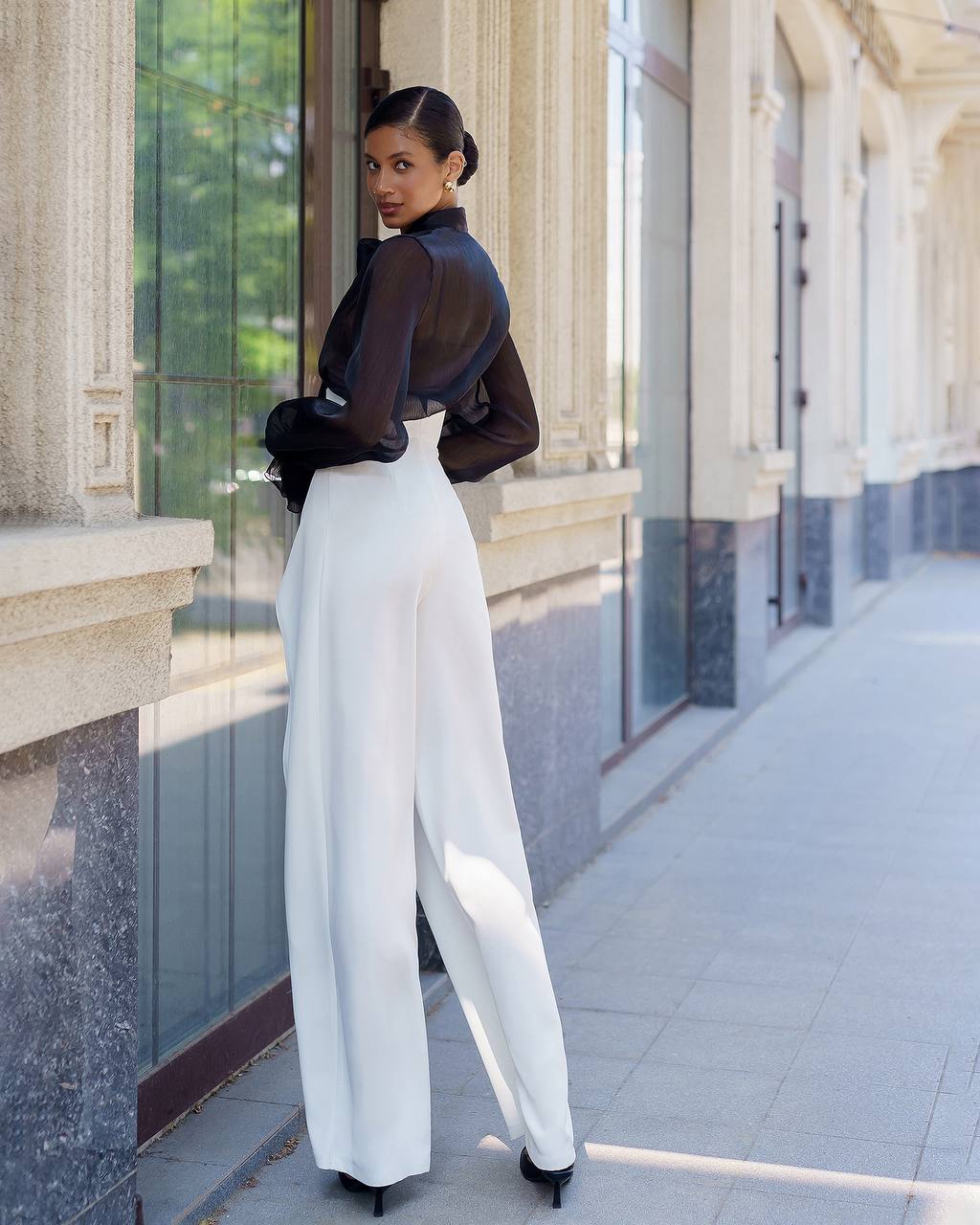 Woman in a black top and white pants standing in front of a building.