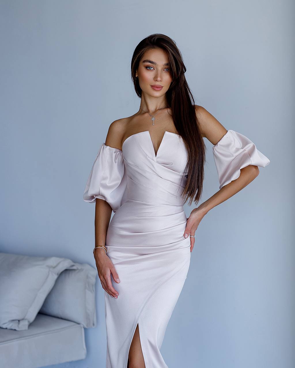 Woman wearing a white off-shoulder dress with a high slit, standing against a light blue wall.