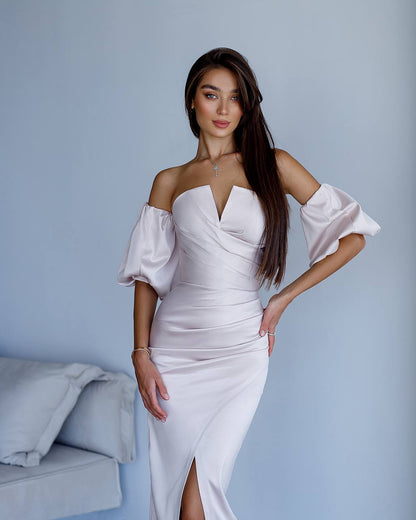 Woman wearing a white off-shoulder dress with a high slit, standing against a light blue wall.