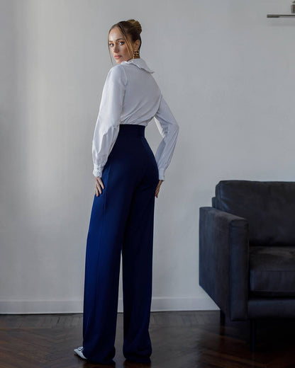 Woman wearing a white blouse and blue pants standing in a room with a gray wall and dark sofa.