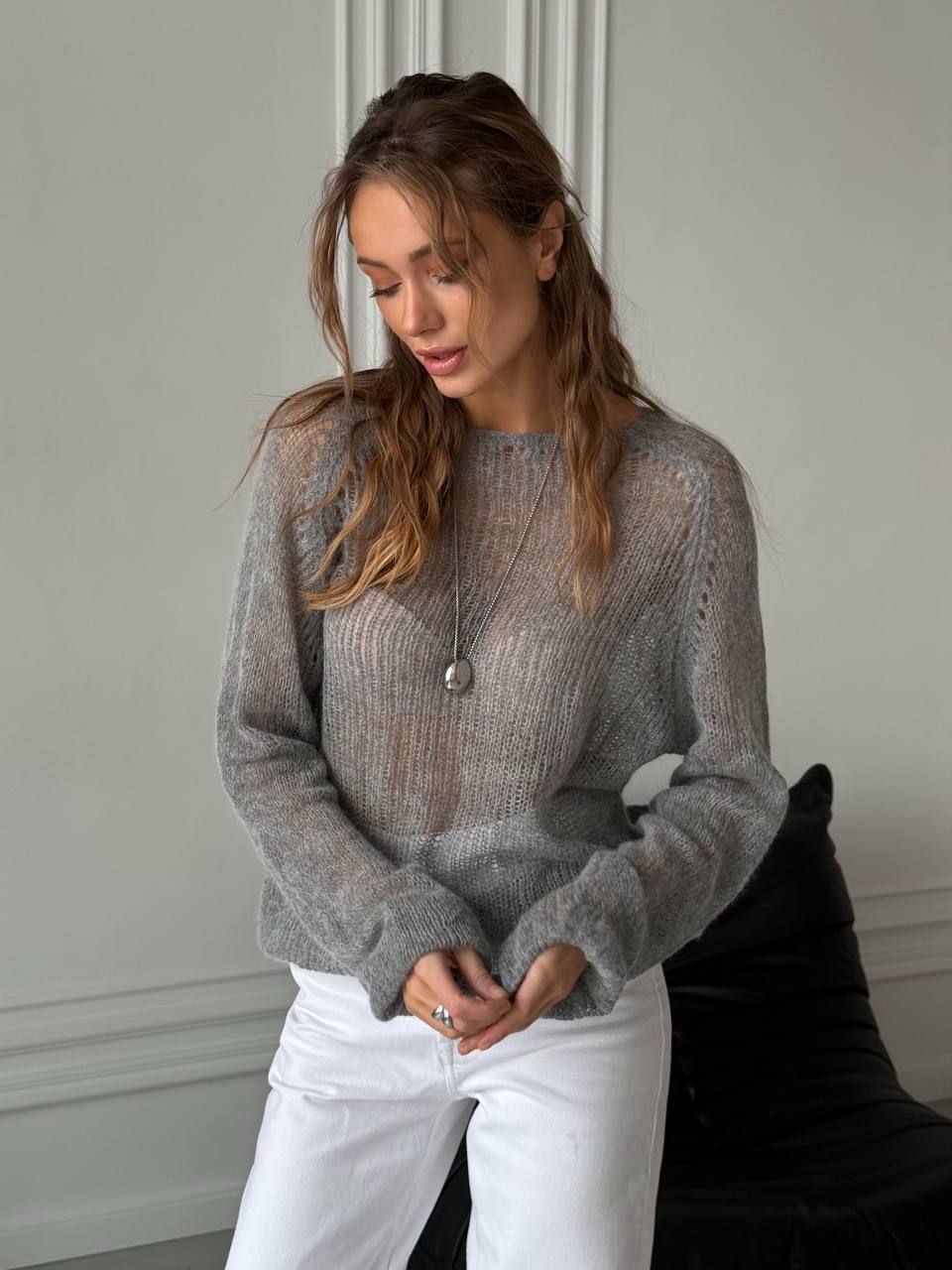Woman wearing a gray sweater and white pants against a neutral background
