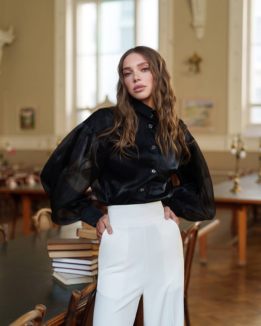 Woman wearing a black blouse and white pants in an indoor setting with large windows.