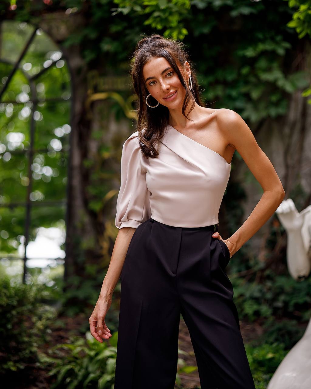 Woman wearing a one-shoulder white top and black pants in a garden setting