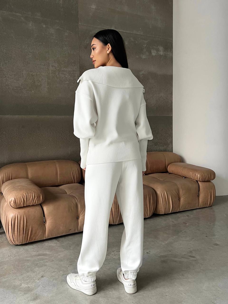 Person wearing a light gray outfit standing in a room with a brown sofa.