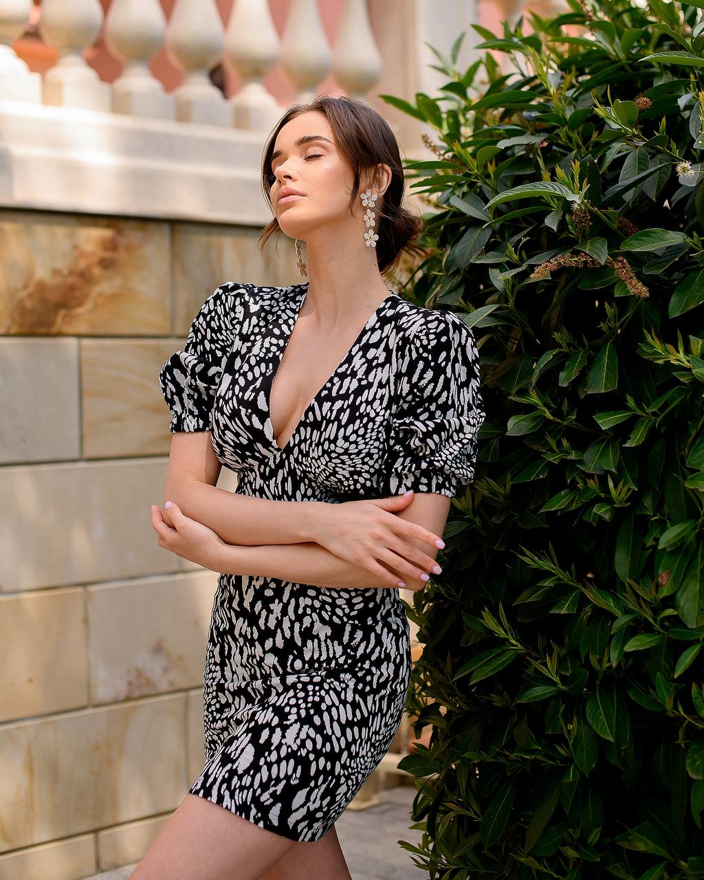 Woman in a black and white patterned dress standing against a stone wall with greenery.
