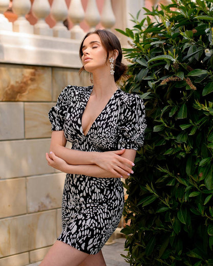 Woman in a black and white patterned dress standing against a stone wall with greenery.