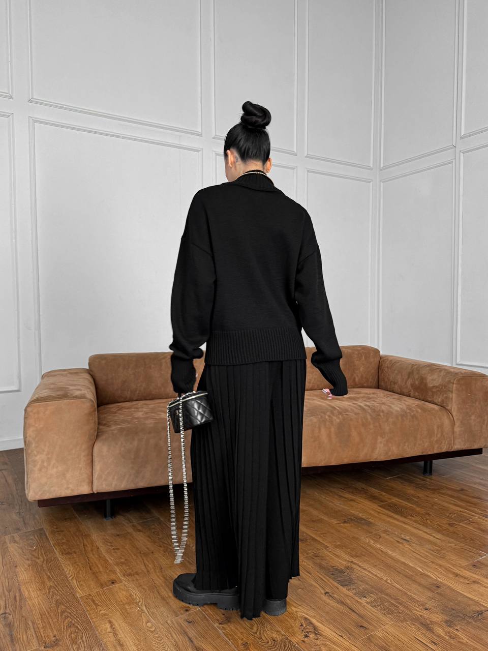 Person in black outfit standing in a room with a brown sofa and white paneled walls.
