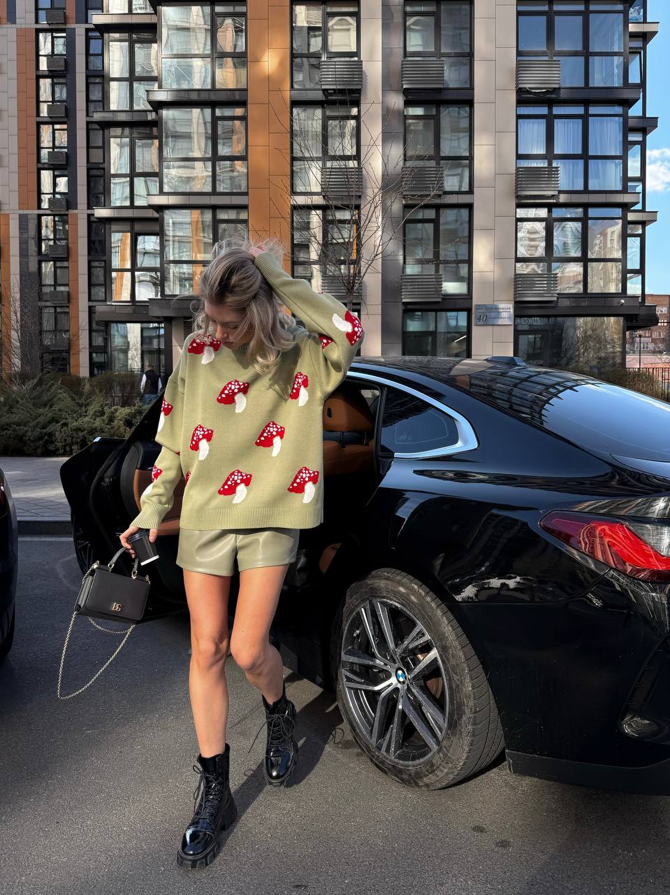 Person wearing a light green sweater with red patterns, black shorts, and black boots standing next to a black car in an urban setting.