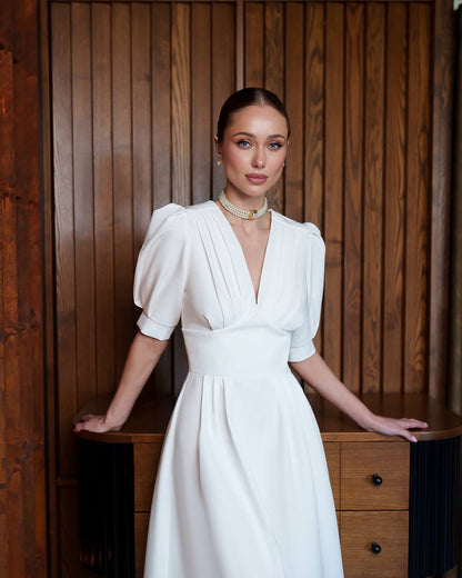 Woman in a white dress standing against a wooden paneled wall.