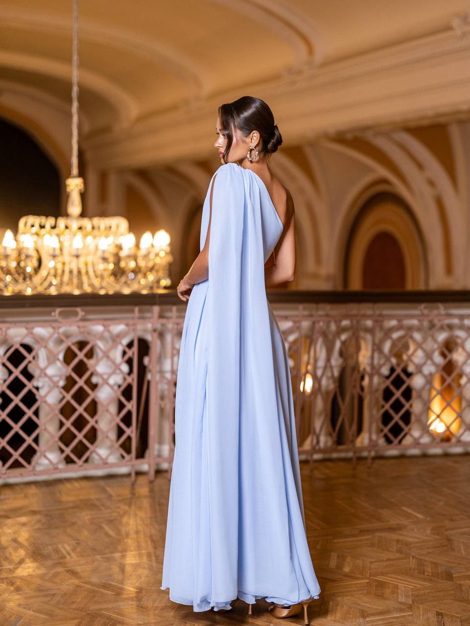 Woman in a light blue evening gown standing in an elegant indoor setting with chandeliers.
