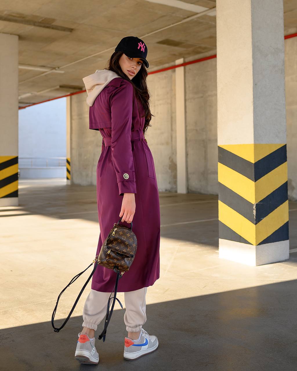Woman in a purple trench coat and white sneakers standing in an industrial setting.