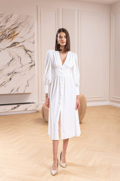 Woman in a white dress standing in a room with marble wall and wooden floor.