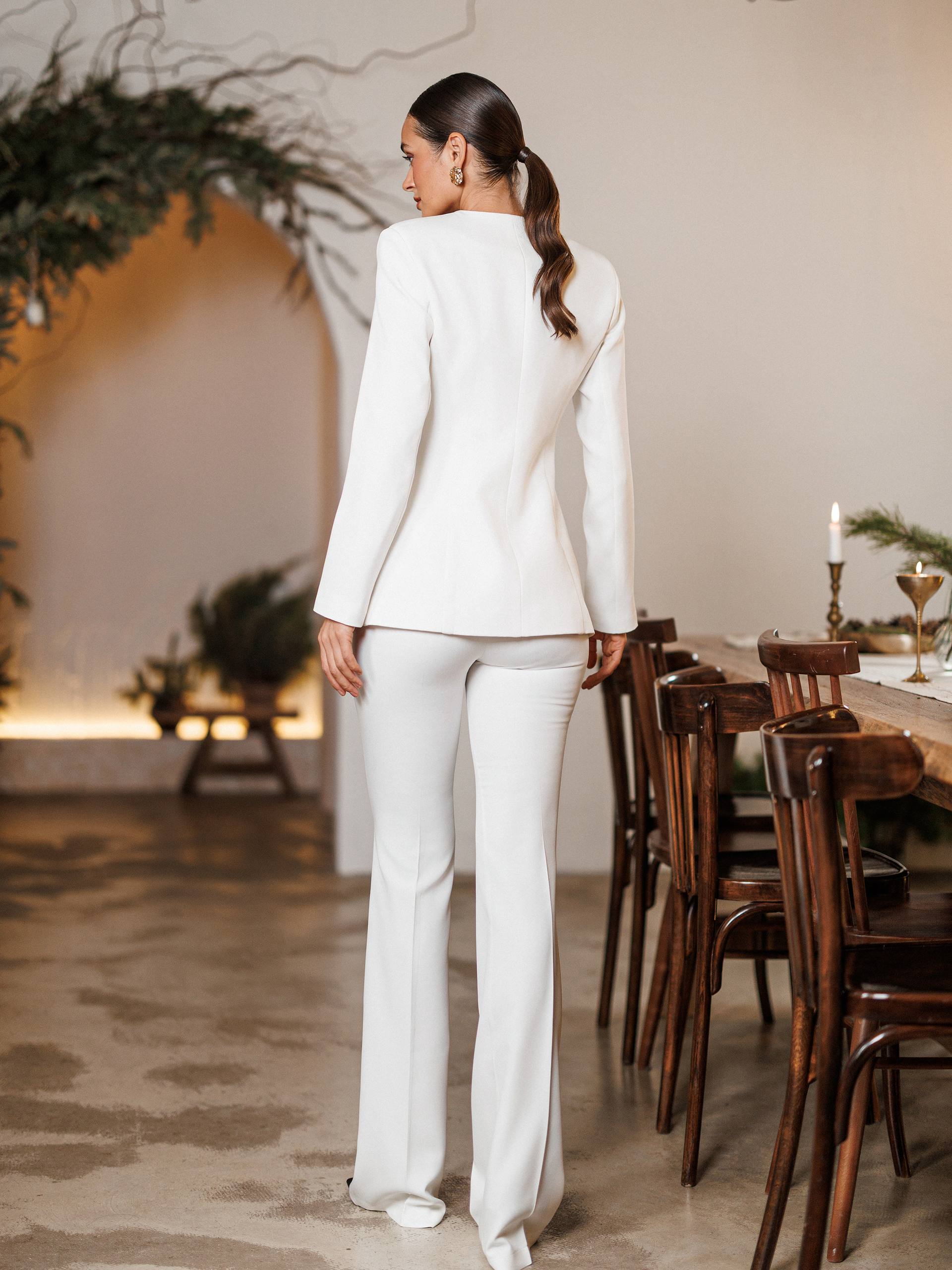 Woman in a white suit standing in a dining room with wooden chairs and a long table.