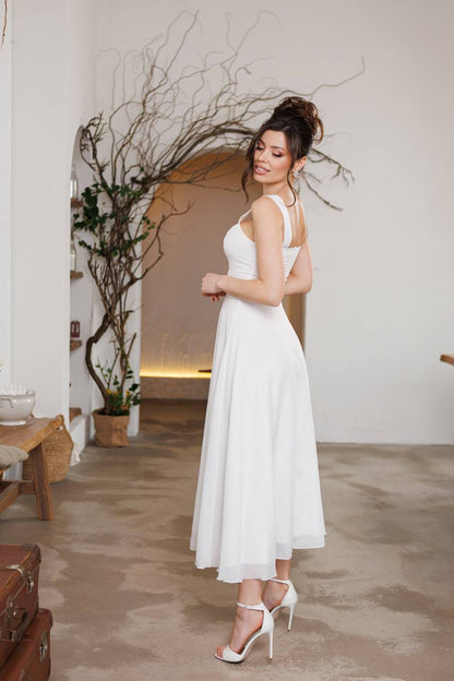 Woman in a white dress standing in an indoor setting with plants and a wooden table.