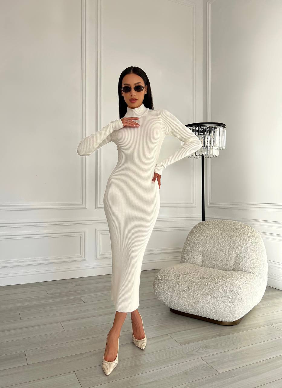 Woman in a white ribbed dress standing in a stylish room with a lamp and chair.