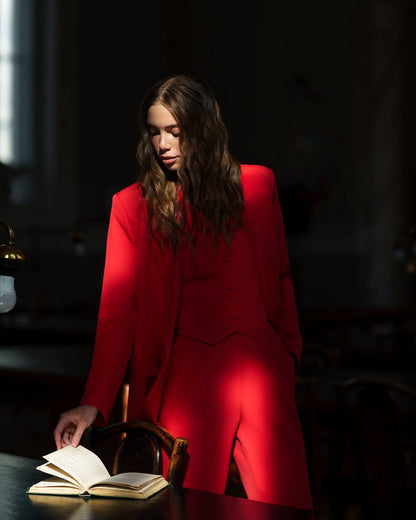 Woman in a red suit sitting at a table with an open book in a dimly lit room.