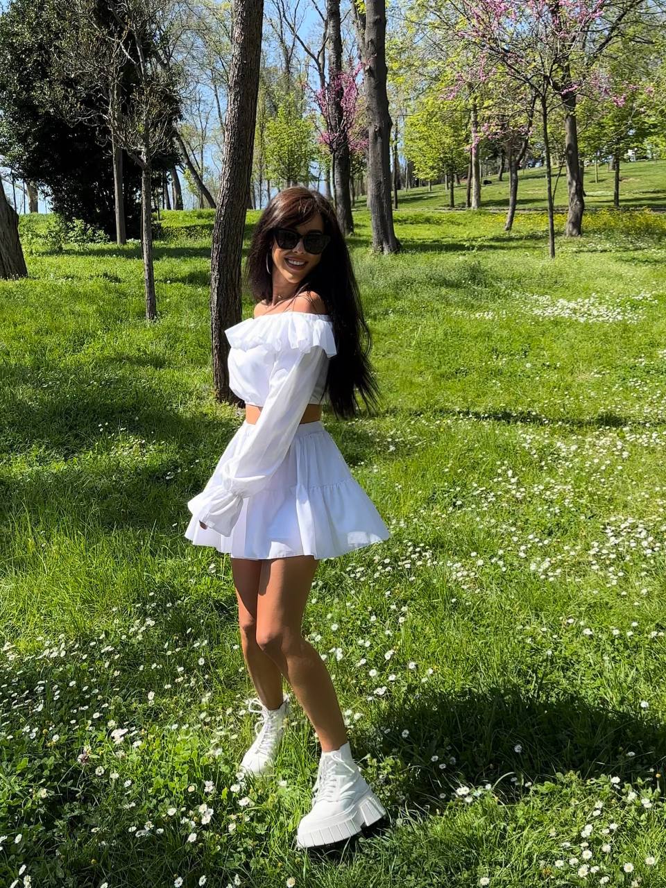 Woman in a white outfit standing in a park with green grass and trees.