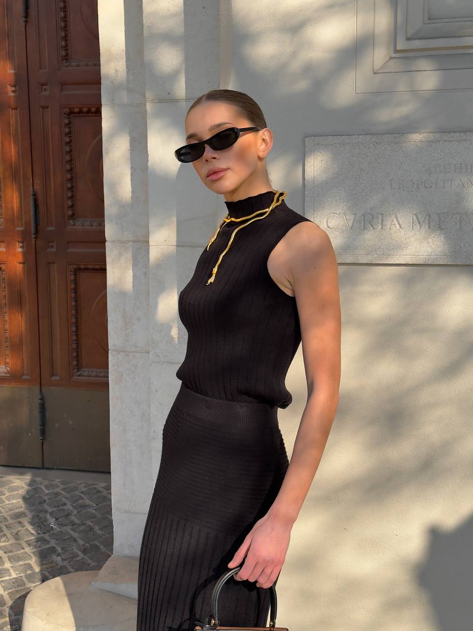 Woman in a black outfit with gold accents standing against a stone wall.