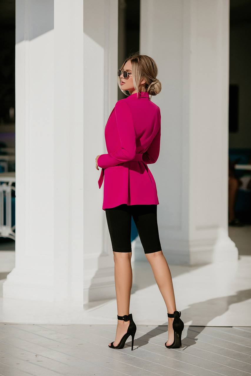 Woman in a bright pink blazer and black shorts standing against white columns.