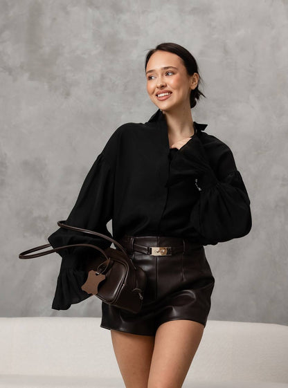 Woman in black outfit with leather shorts and bag against a gray background