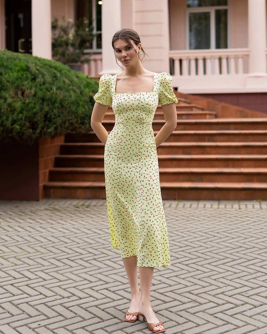 Woman in a light green floral dress standing on a paved area with steps and greenery in the background.