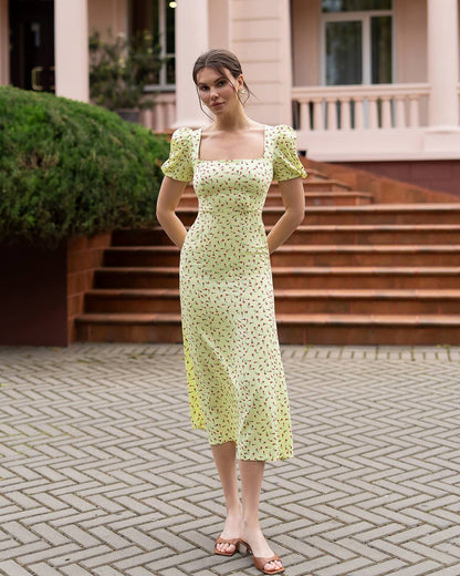 Woman in a light green floral dress standing on a paved area with steps and greenery in the background.