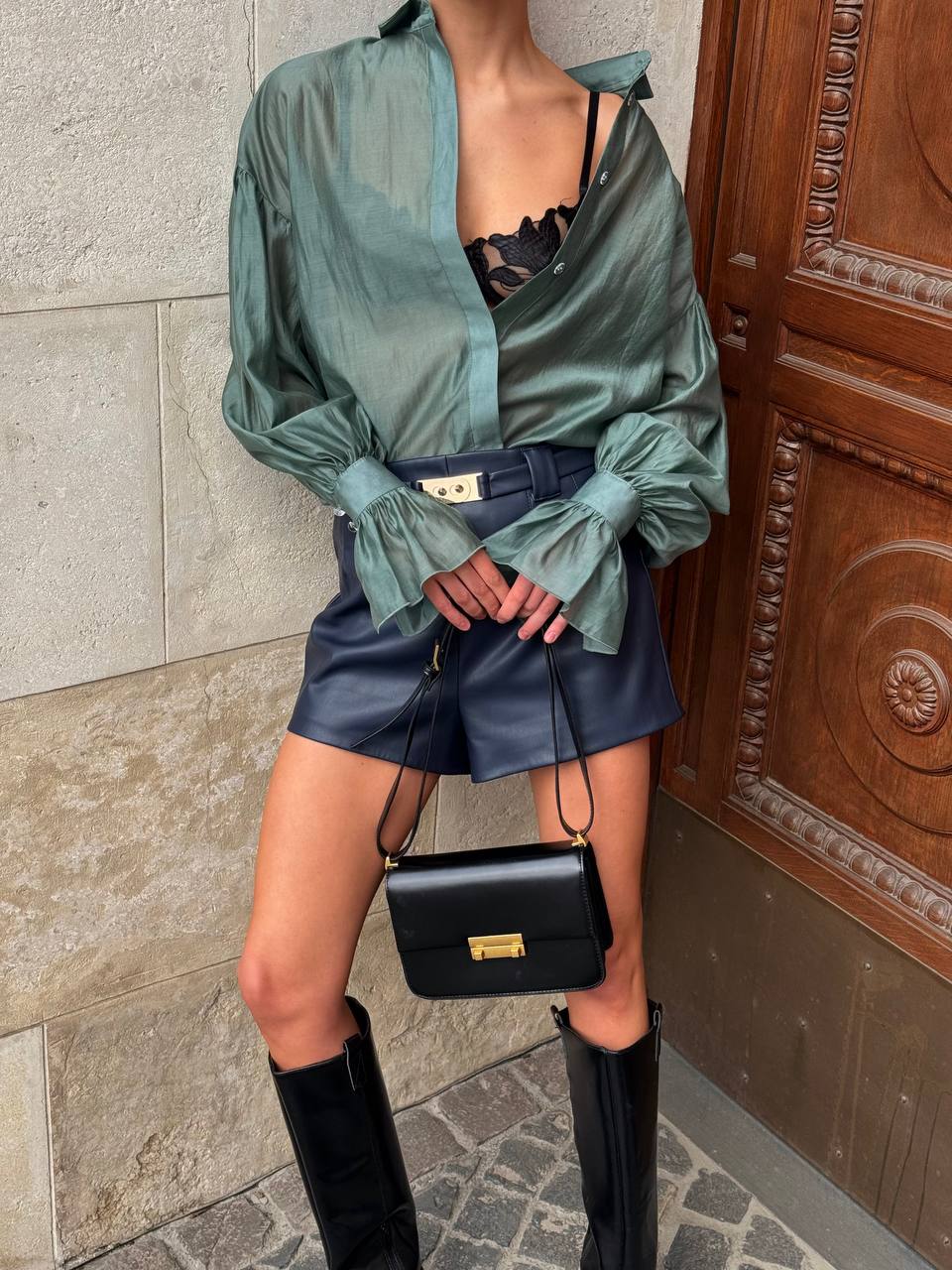 Person wearing a green satin blouse, black leather skirt, and knee-high boots, holding a black handbag.