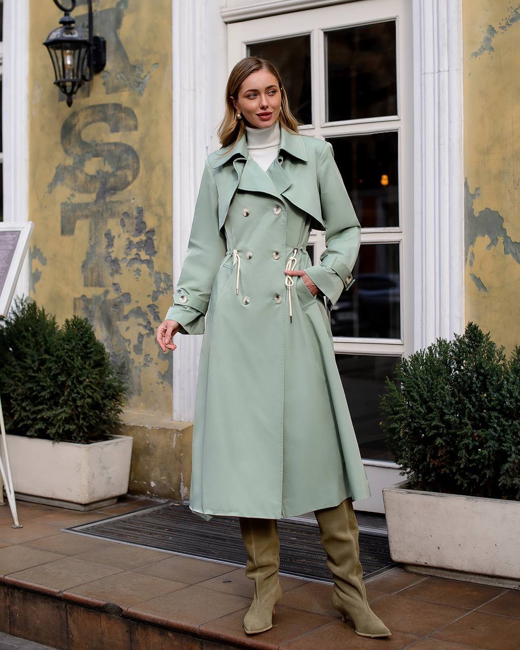 Woman wearing a light green trench coat standing outdoors with a building and plants in the background.